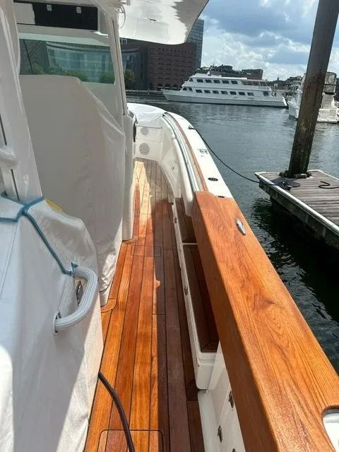 The side deck of a boat featuring teak flooring and wood gunwales, looking out toward a marina and a docked cruise ship.