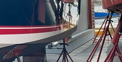 A boat hull painted red, white, and black supported by metal stands in a boatyard.