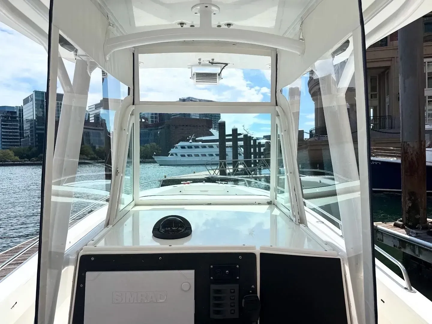 View from the helm of a boat looking past a windshield and canvas enclosure toward a harbor with buildings and a yacht.