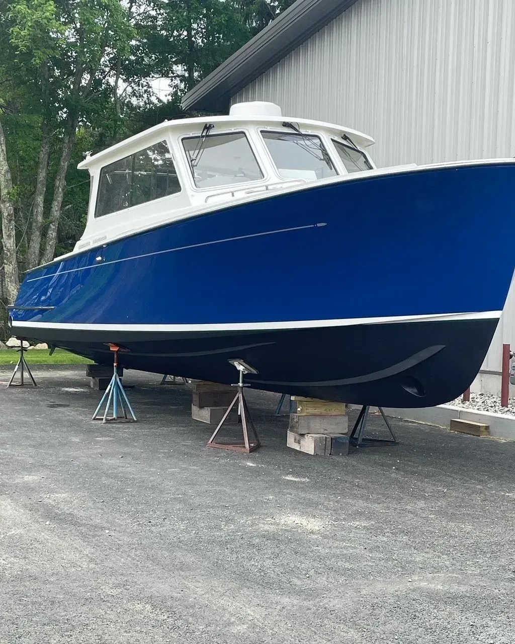 A blue and white motorboat resting on metal jack stands in a gravel lot next to a white building.