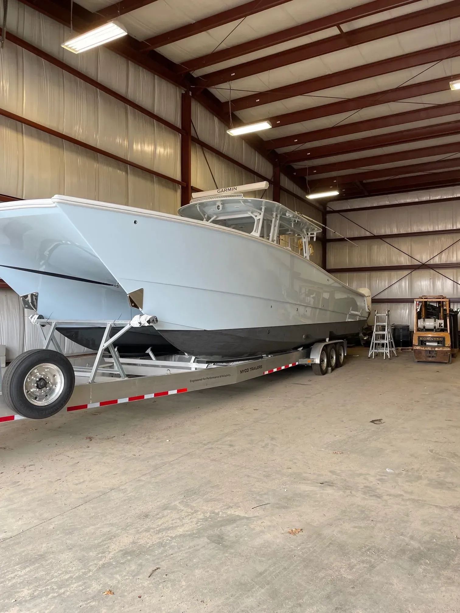 A large, light-gray catamaran boat on a multi-axle trailer inside a metal warehouse.