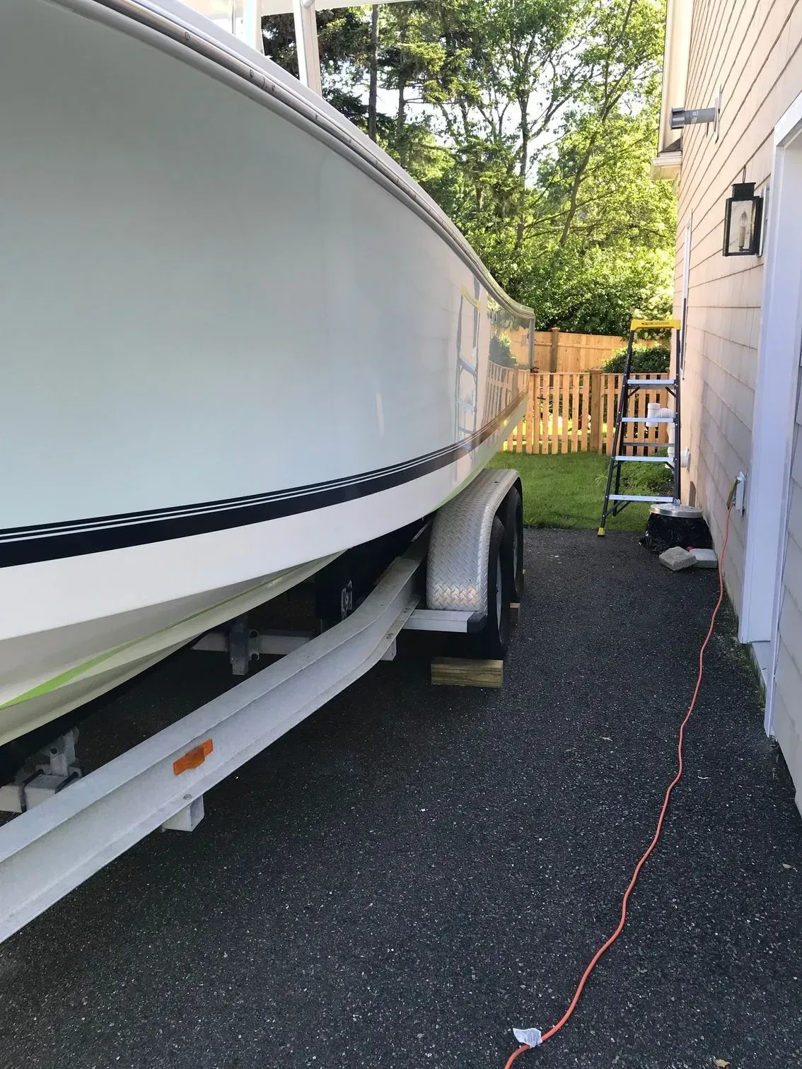 A light blue boat on a trailer parked on a gravel driveway beside a house.