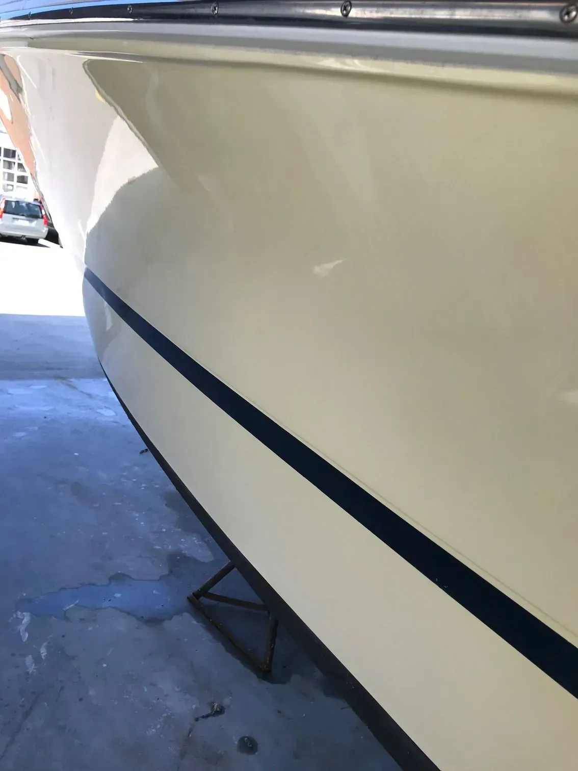 Side view of a light-colored boat hull with a black stripe, resting on a stand on a concrete surface.