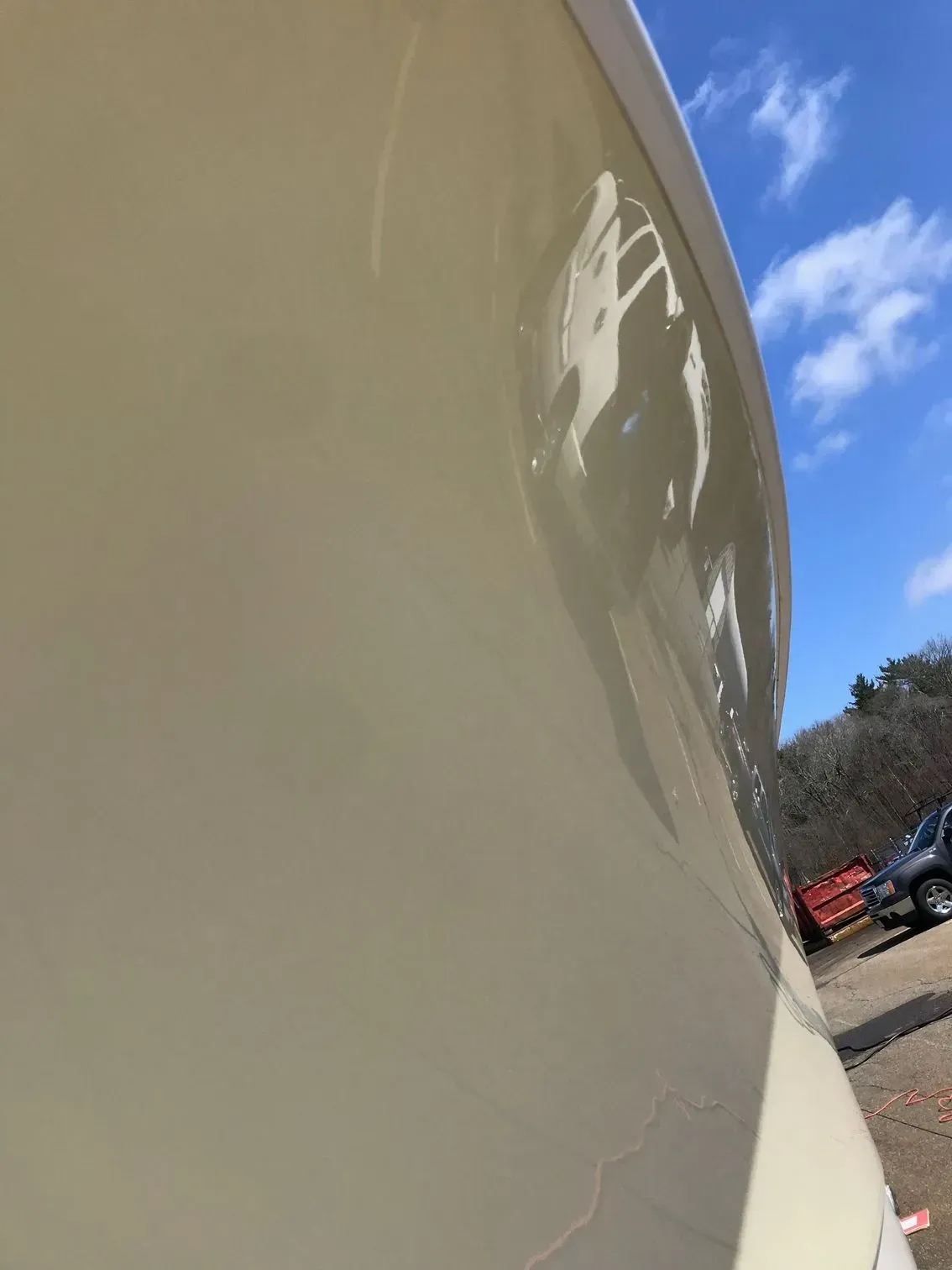 A close-up, angled view of a smooth, beige boat hull reflecting the blue sky and a vehicle in an outdoor lot.