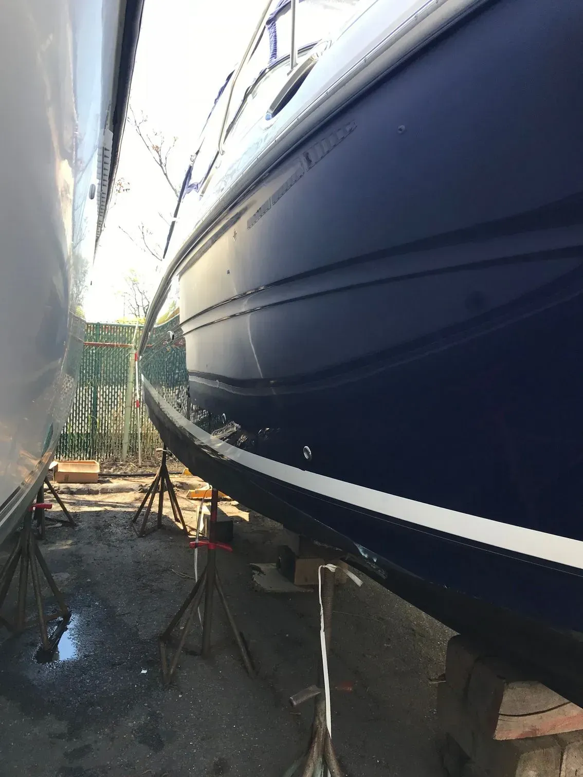 A blue boat sits on metal stands in an outdoor storage yard, next to the hull of another boat.