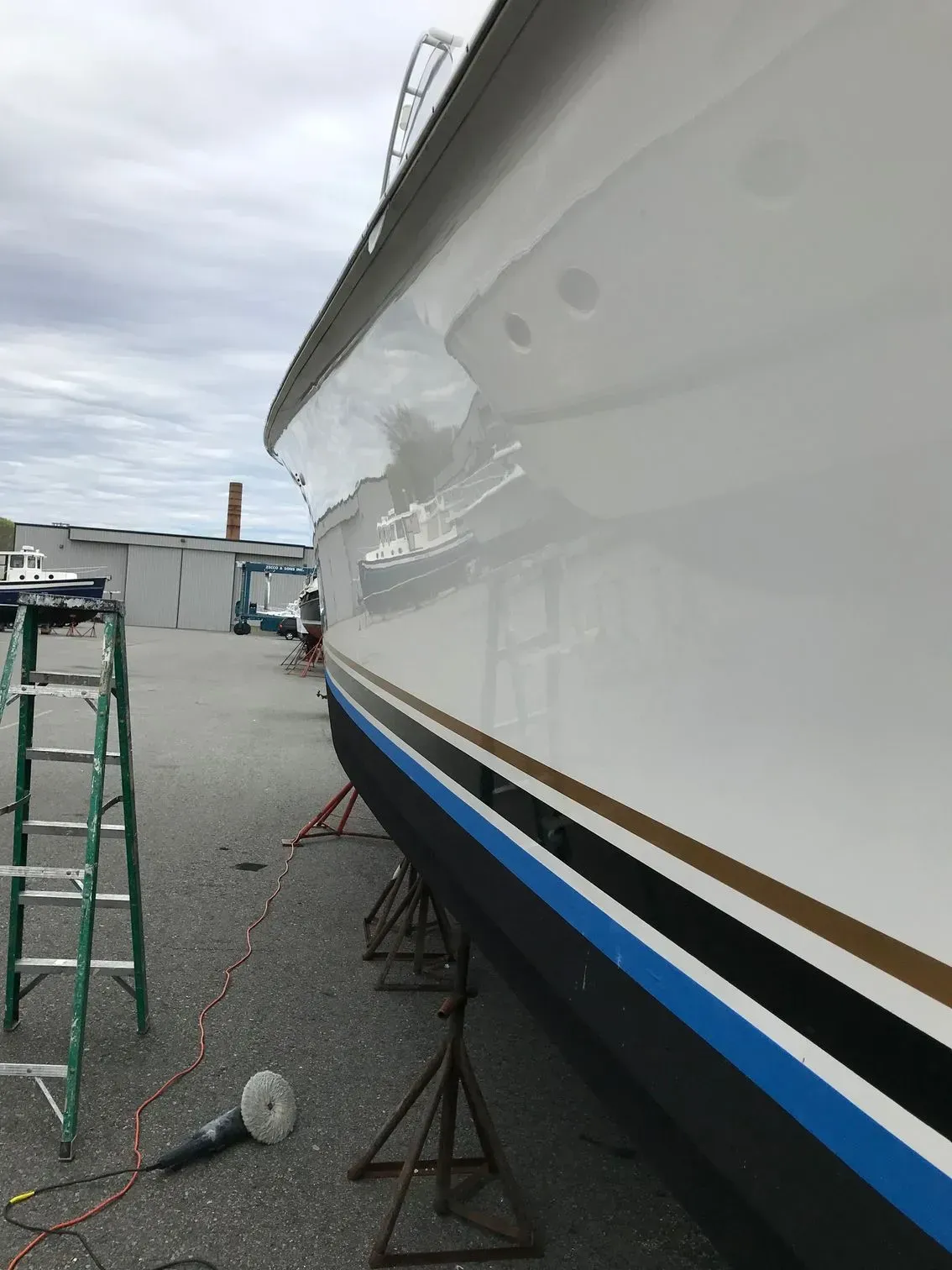 A white boat hull in a shipyard, propped up by metal stands, with a green ladder and a power polisher nearby.