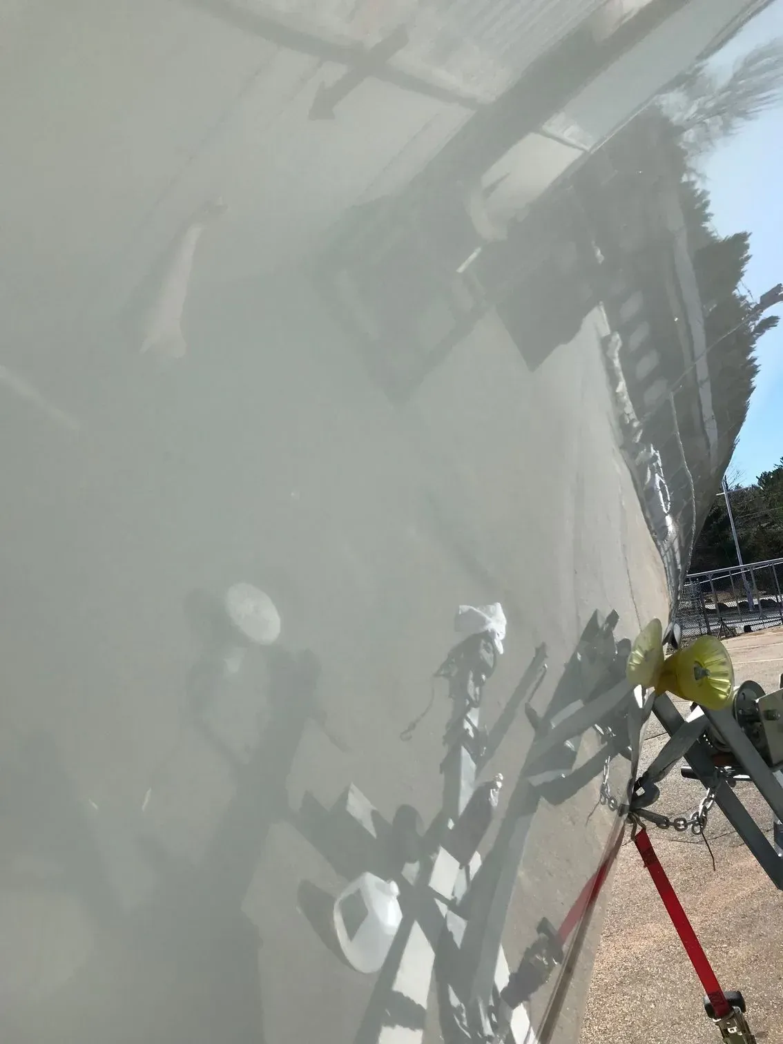 A close-up of a glossy, light-colored boat hull reflecting its trailer, the ground, and surrounding trees.