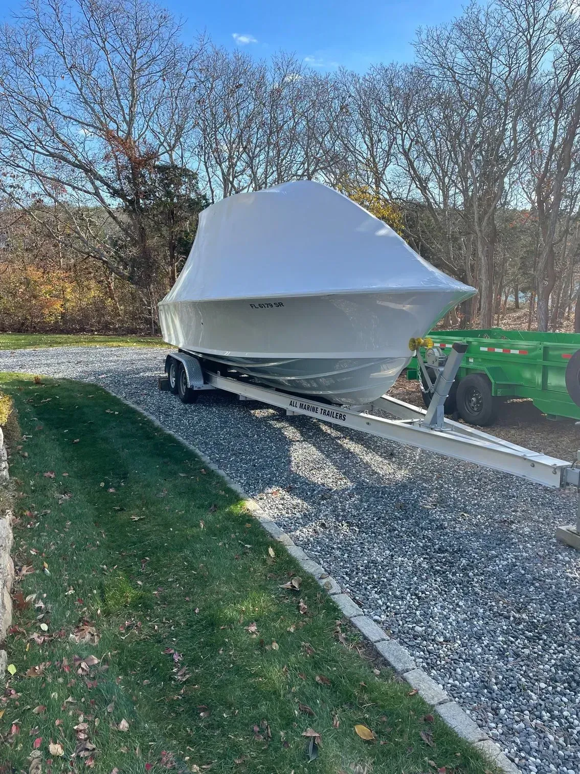 A boat covered in a white shrink-wrap tarp on a trailer, parked on a gravel driveway beside a grassy lawn.