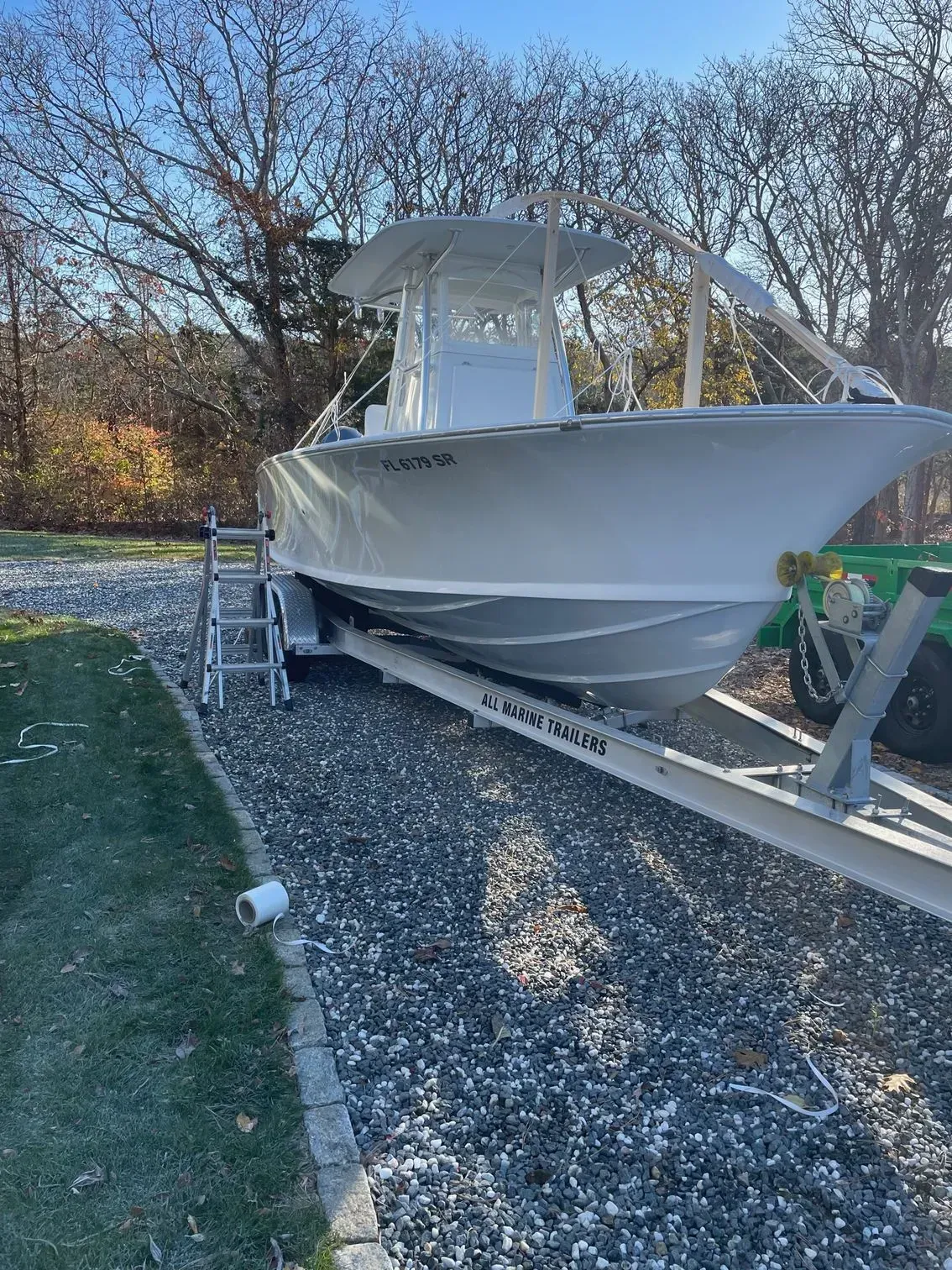 A white center console boat on a trailer parked on a gravel driveway near a grassy lawn and trees.