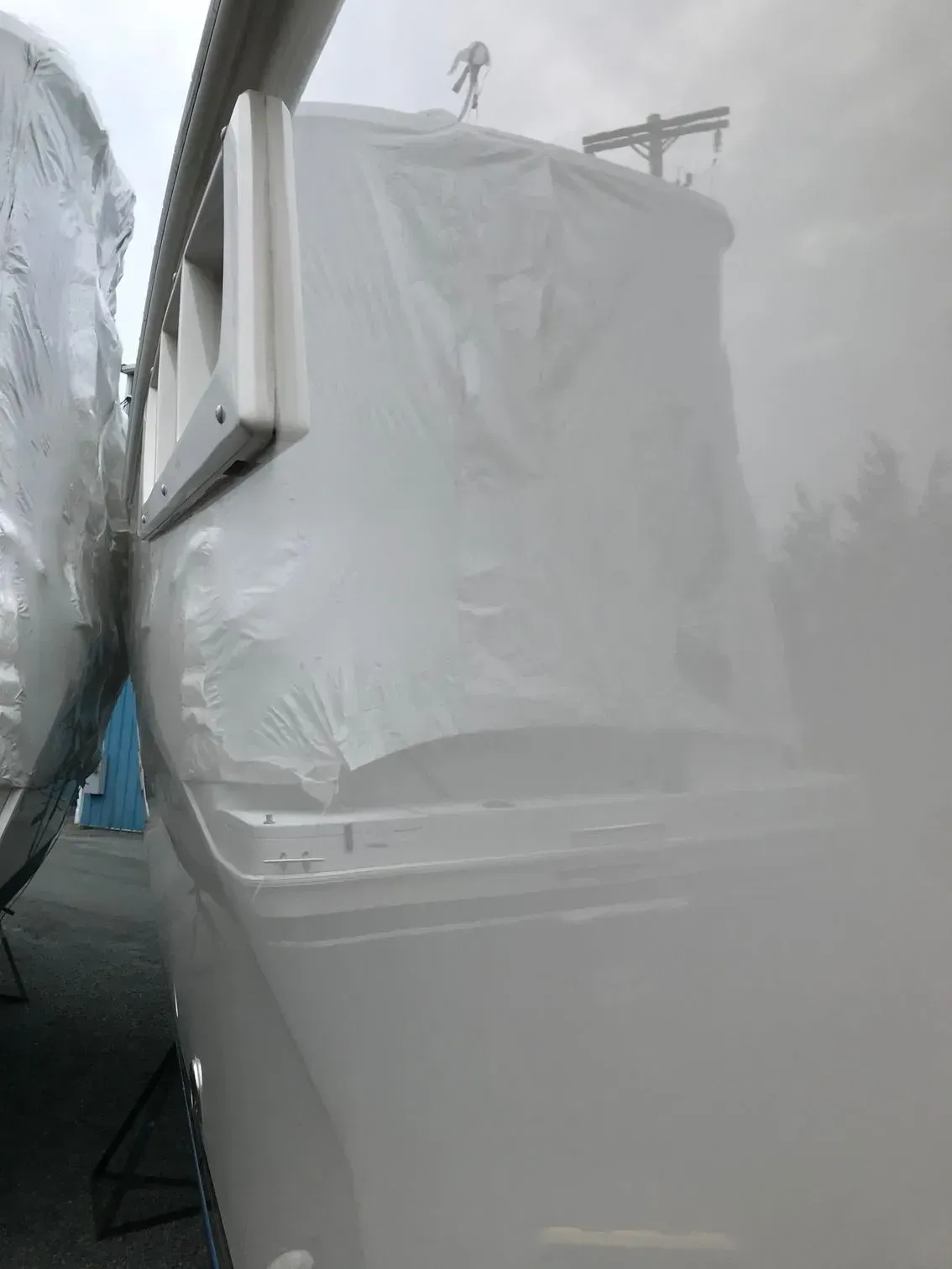 A boat hull wrapped in white protective plastic film stands in an outdoor storage yard.