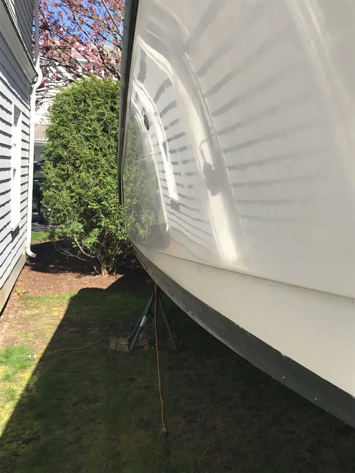 Side view of a white boat hull resting on a metal stand in a grassy yard next to a house.