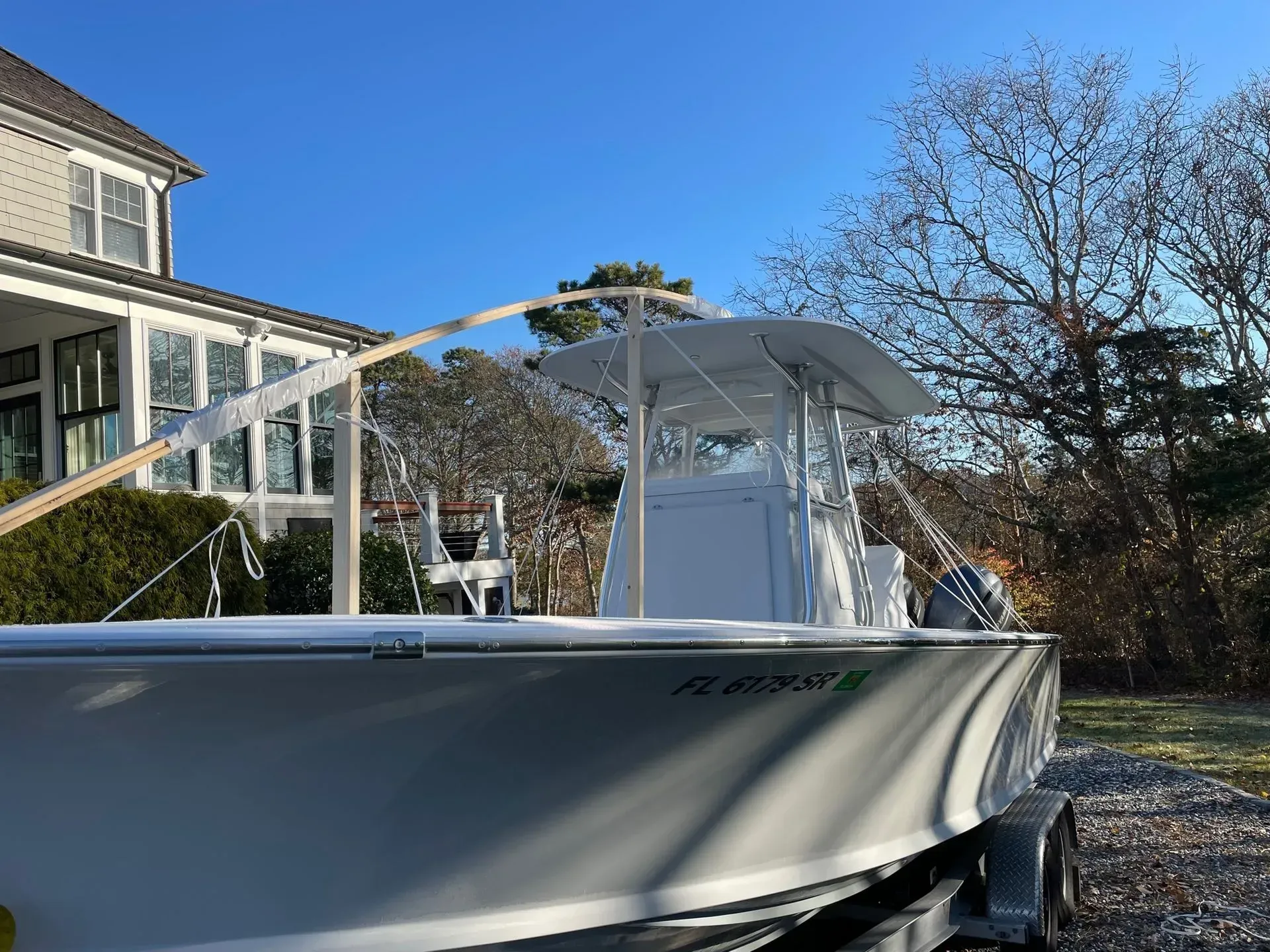 A white center console boat on a trailer, parked on a gravel surface in front of a house on a sunny day.