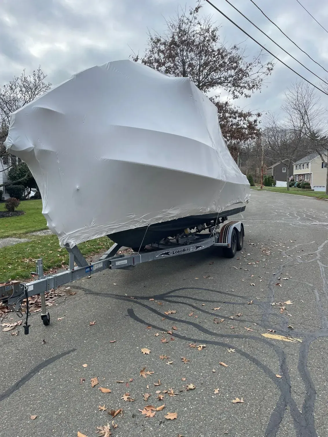 A boat wrapped in white protective shrink wrap, sitting on a trailer on a residential street during autumn.