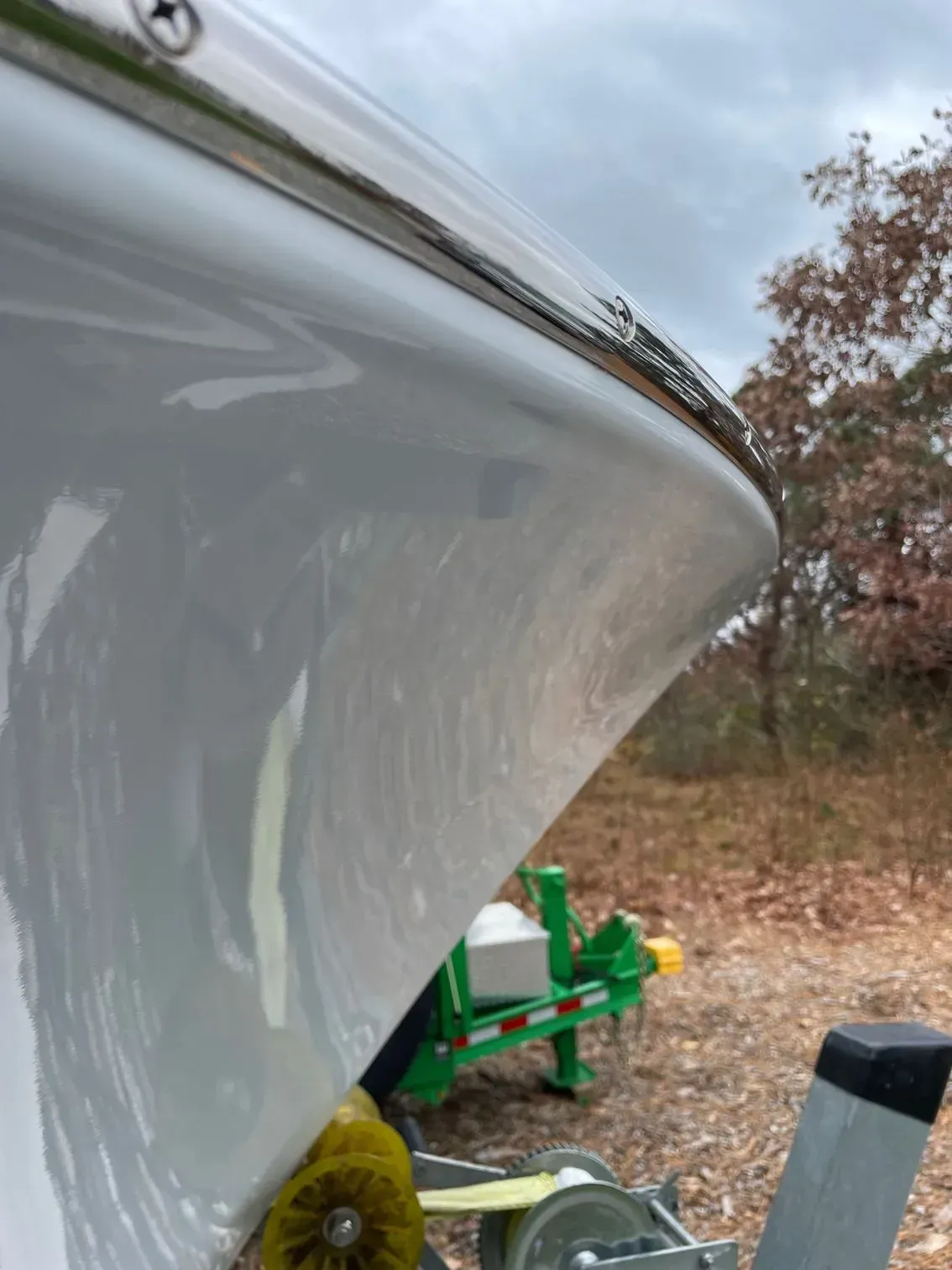 Close-up of a white boat hull on a trailer, showing a damaged metal rub rail against an outdoor, wooded background.
