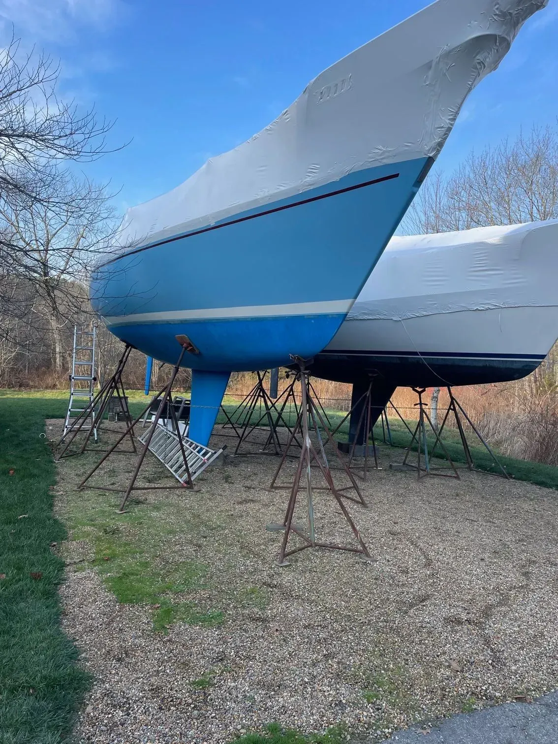 Two sailboats in winter storage on gravel, propped up by metal jack stands and covered in protective white plastic wrap.
