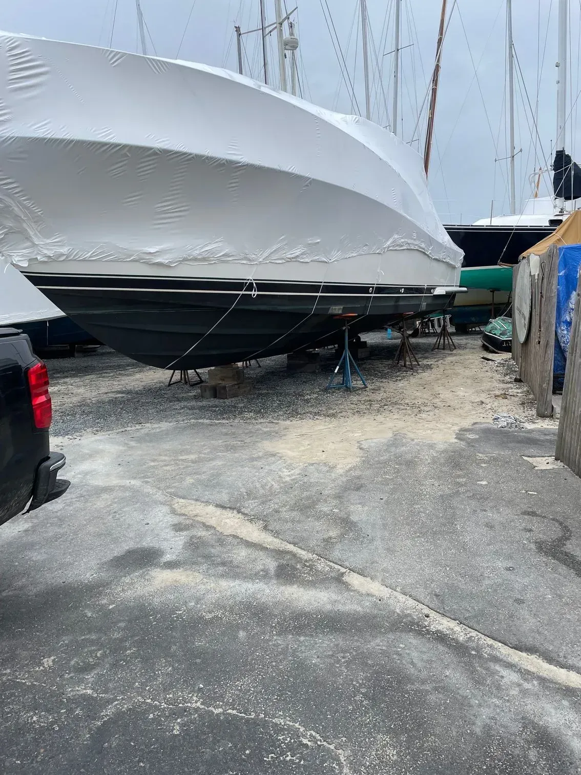 A boat in a shipyard, covered in white shrink wrap, supported by metal stands on a gravel lot next to a vehicle.