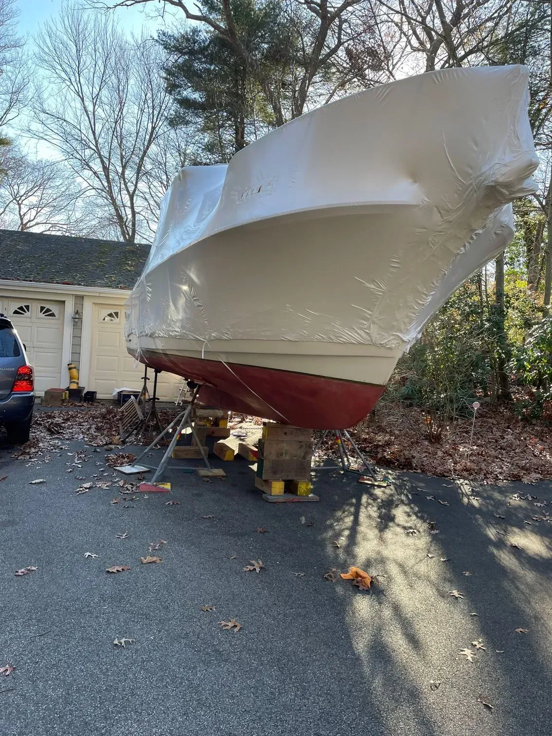 A white shrink-wrapped boat on wooden blocks sits in a driveway in front of a garage surrounded by bare autumn trees.