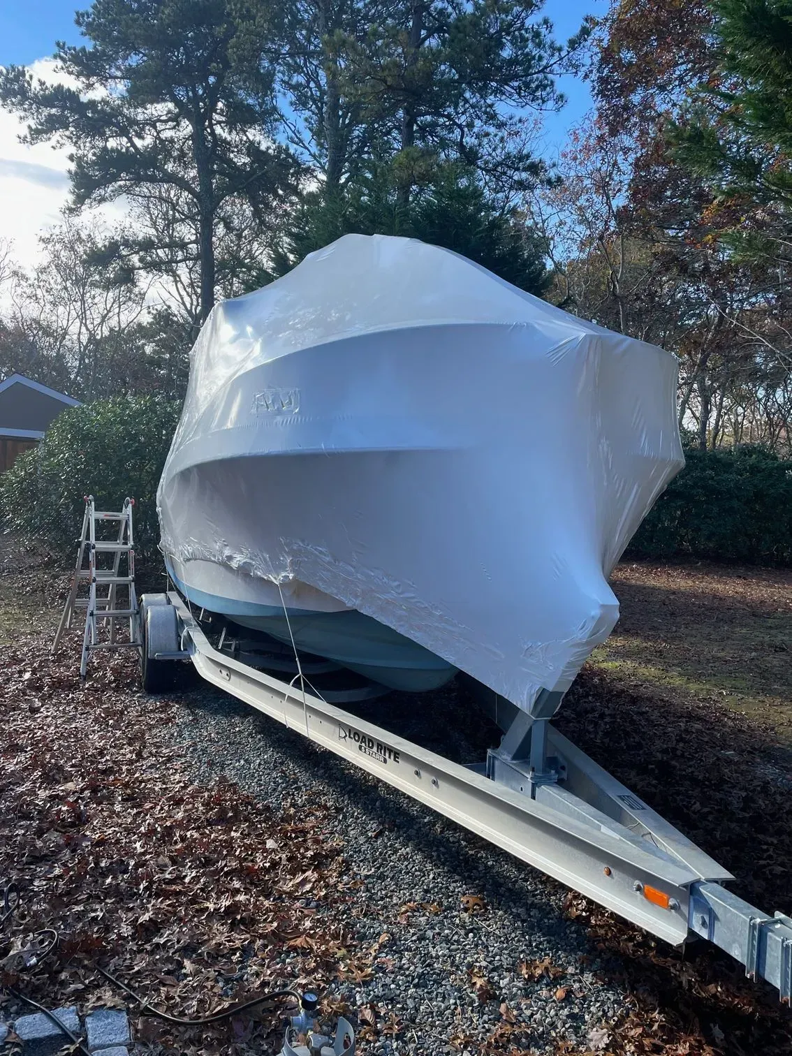 A boat wrapped in white protective plastic sheeting sits on a trailer in a wooded area covered with autumn leaves.