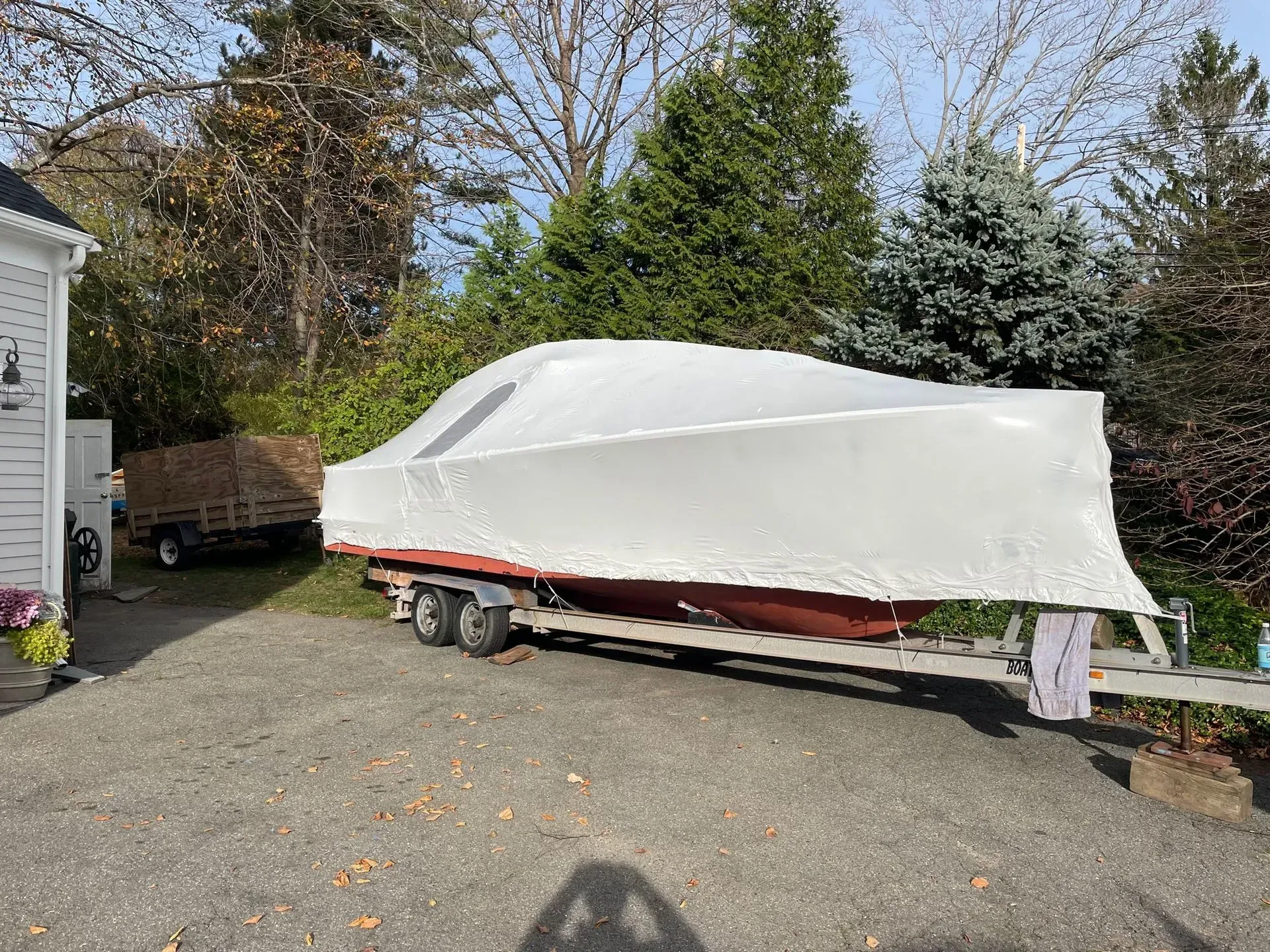 A boat wrapped in white plastic shrink wrap on a trailer parked on a gravel driveway next to a house.