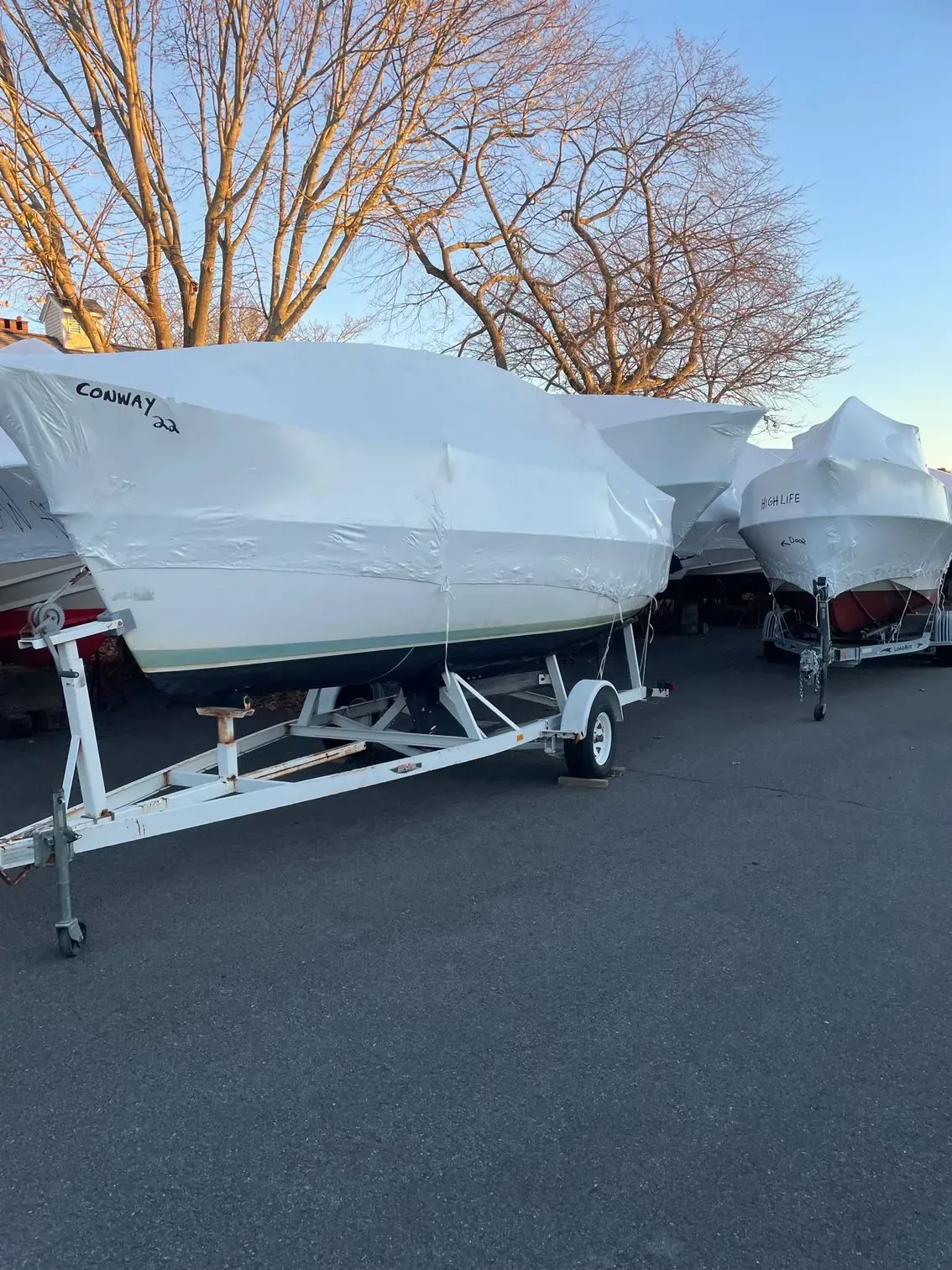 Several boats wrapped in white shrink-wrap on boat trailers in an outdoor lot, set against a backdrop of bare trees.