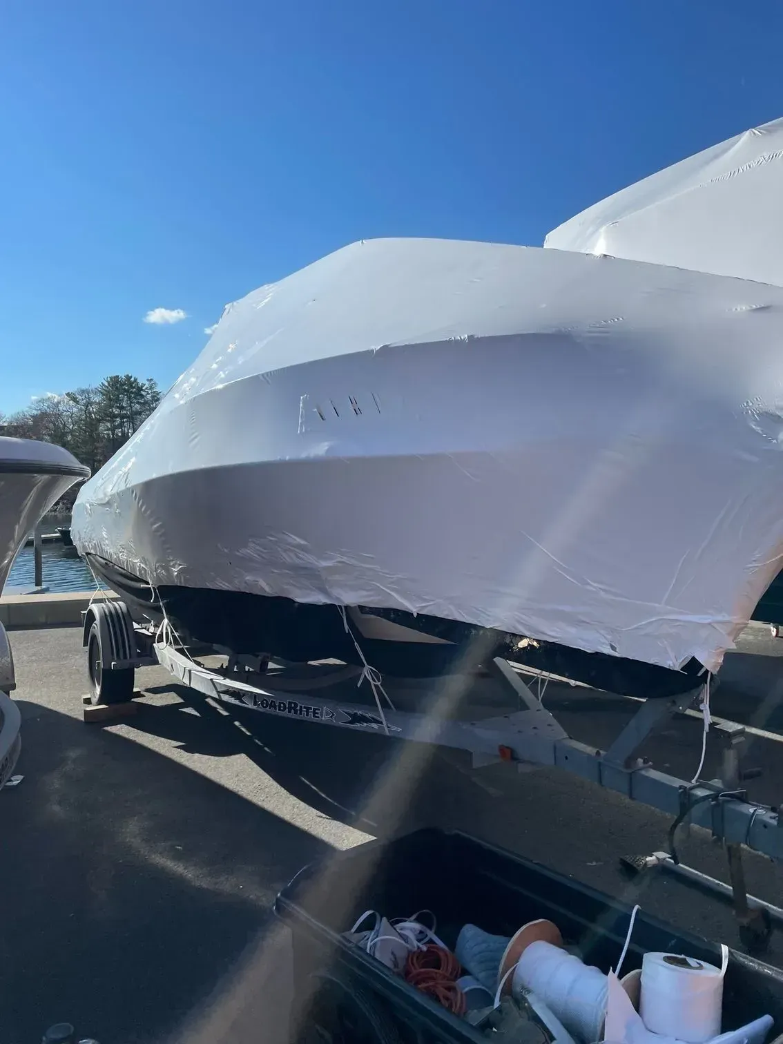 A white shrink-wrapped motorboat secured to a metal trailer in a sunny outdoor lot.