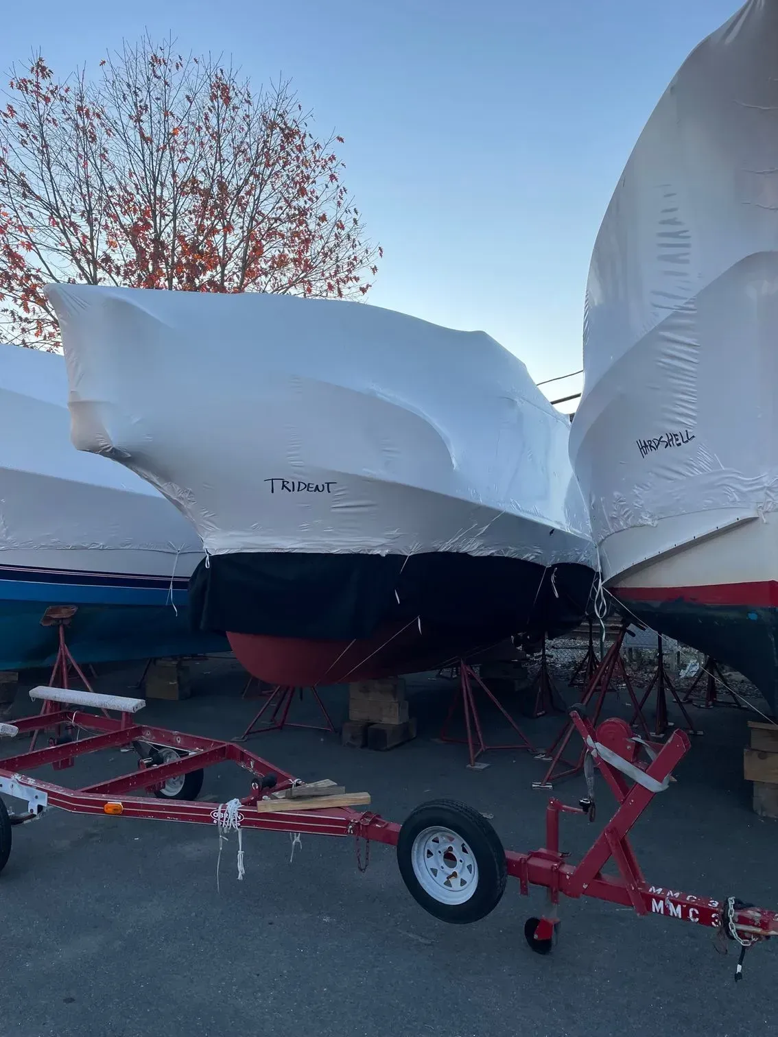 Shrink-wrapped boats in winter storage sit on metal stands in an outdoor lot, with a red boat trailer in the foreground.