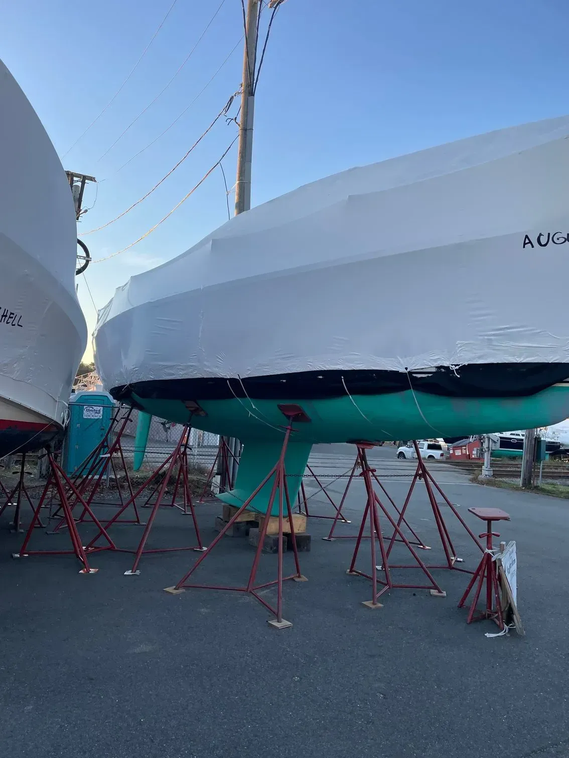 A teal-hulled sailboat on metal supports in a boatyard, covered in white shrink wrap for winter storage.
