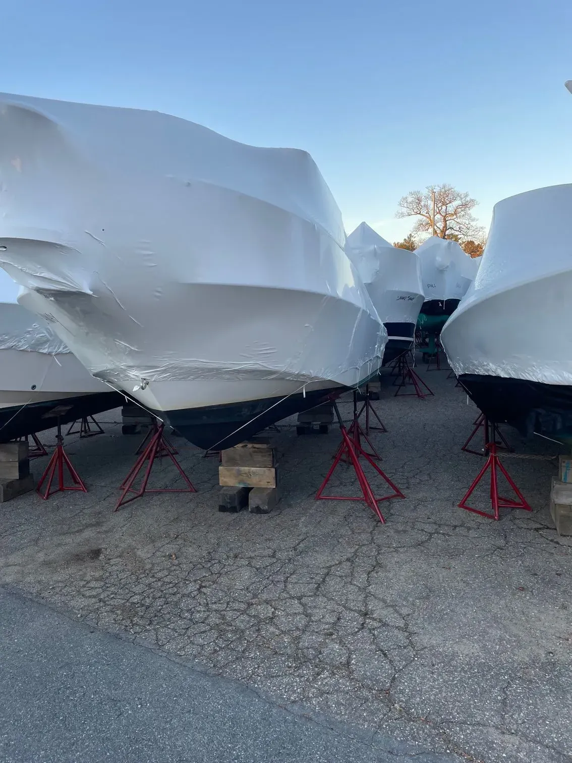 Several boats in a storage yard, each wrapped in white shrink wrap and elevated on red metal stands.