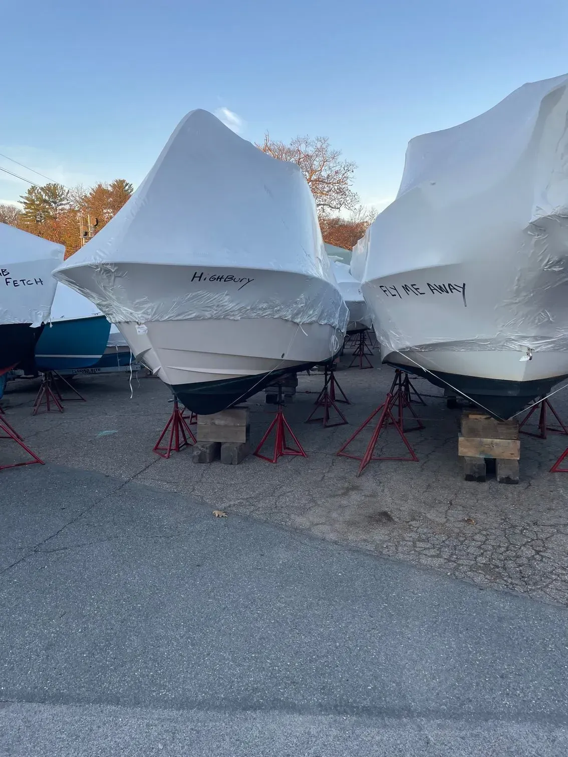 Three boats wrapped in white protective plastic covers sitting on stands in a parking lot during autumn.