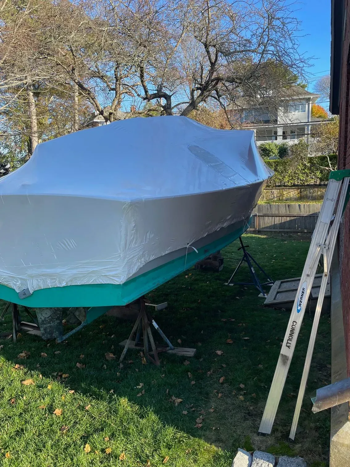 A boat covered in protective shrink wrap sits on stands in a grassy yard, with a ladder leaning against a wall nearby.
