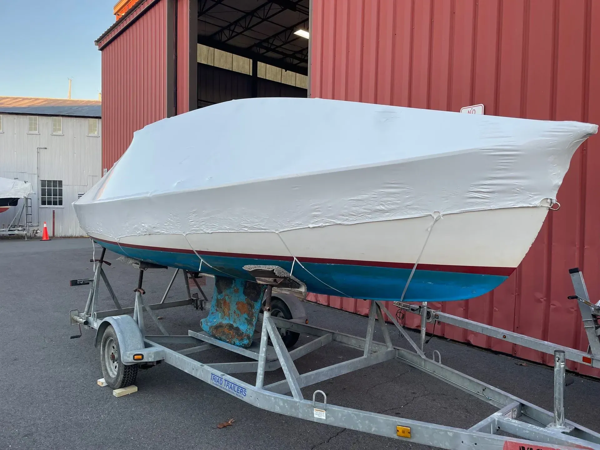 A white sailboat with a blue hull, secured and covered in shrink wrap on a metal trailer parked outside a building.