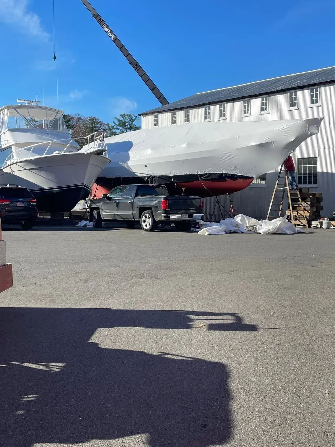 A large boat covered in white shrink-wrap is parked next to a building while being worked on by a person on a ladder.