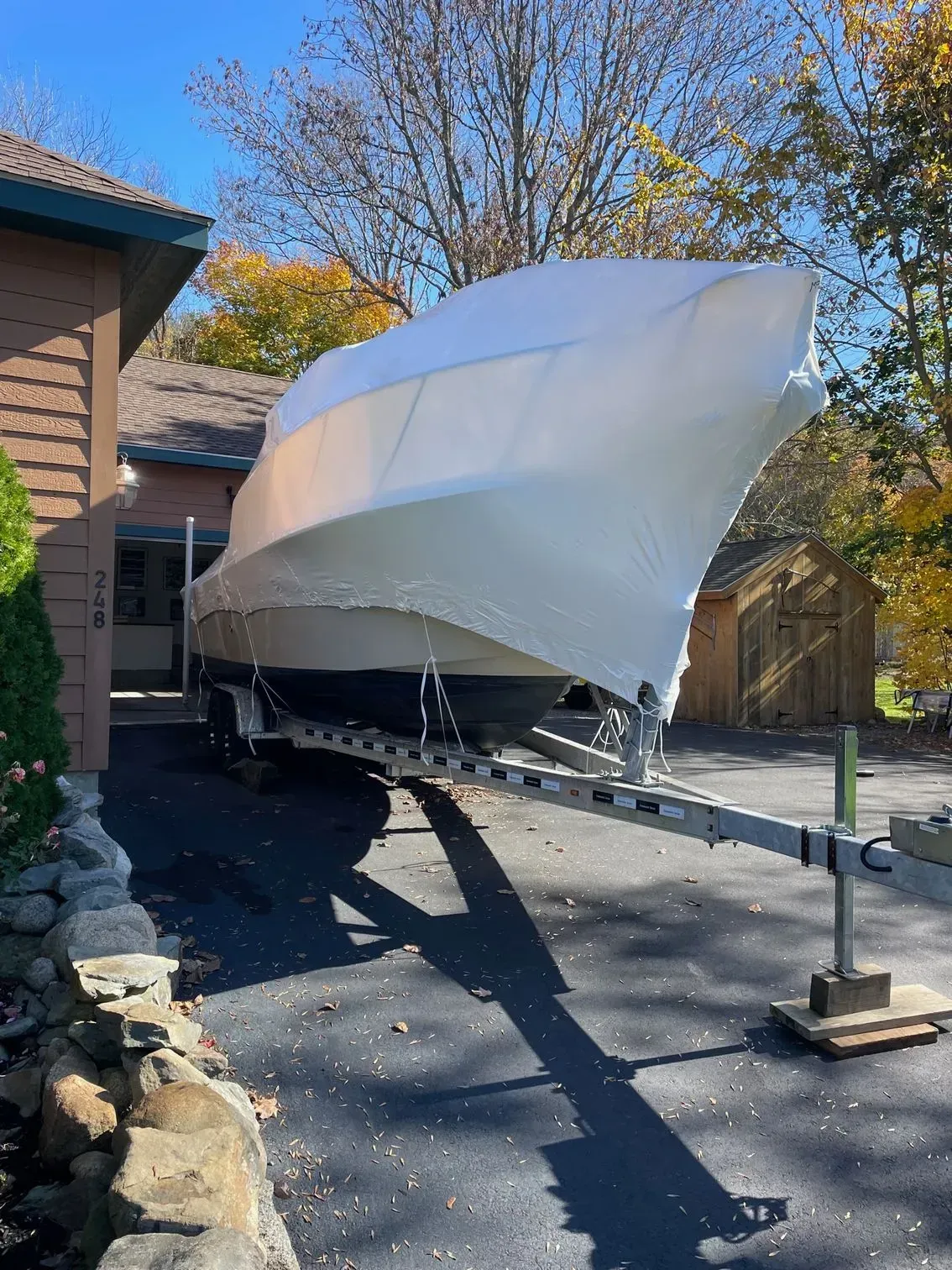 A boat covered in protective white shrink wrap sits on a trailer in a driveway next to a house on a sunny day.
