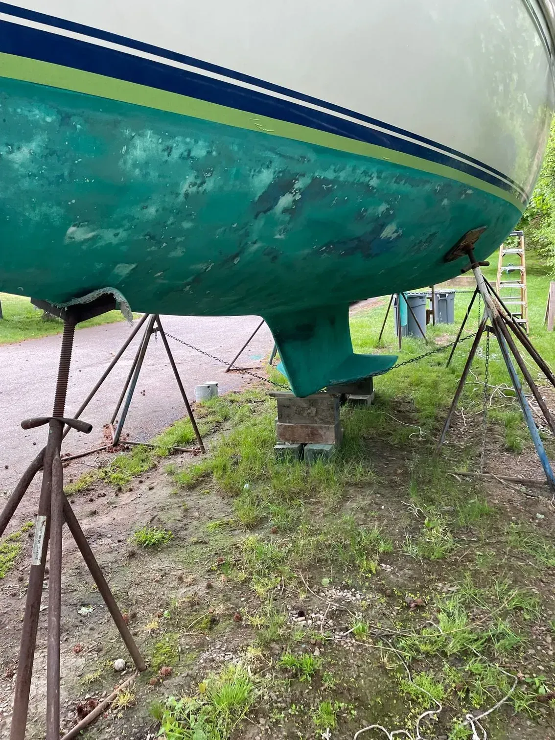 A white boat hull with green bottom paint sits on metal stands and wooden blocks in a grassy outdoor area.