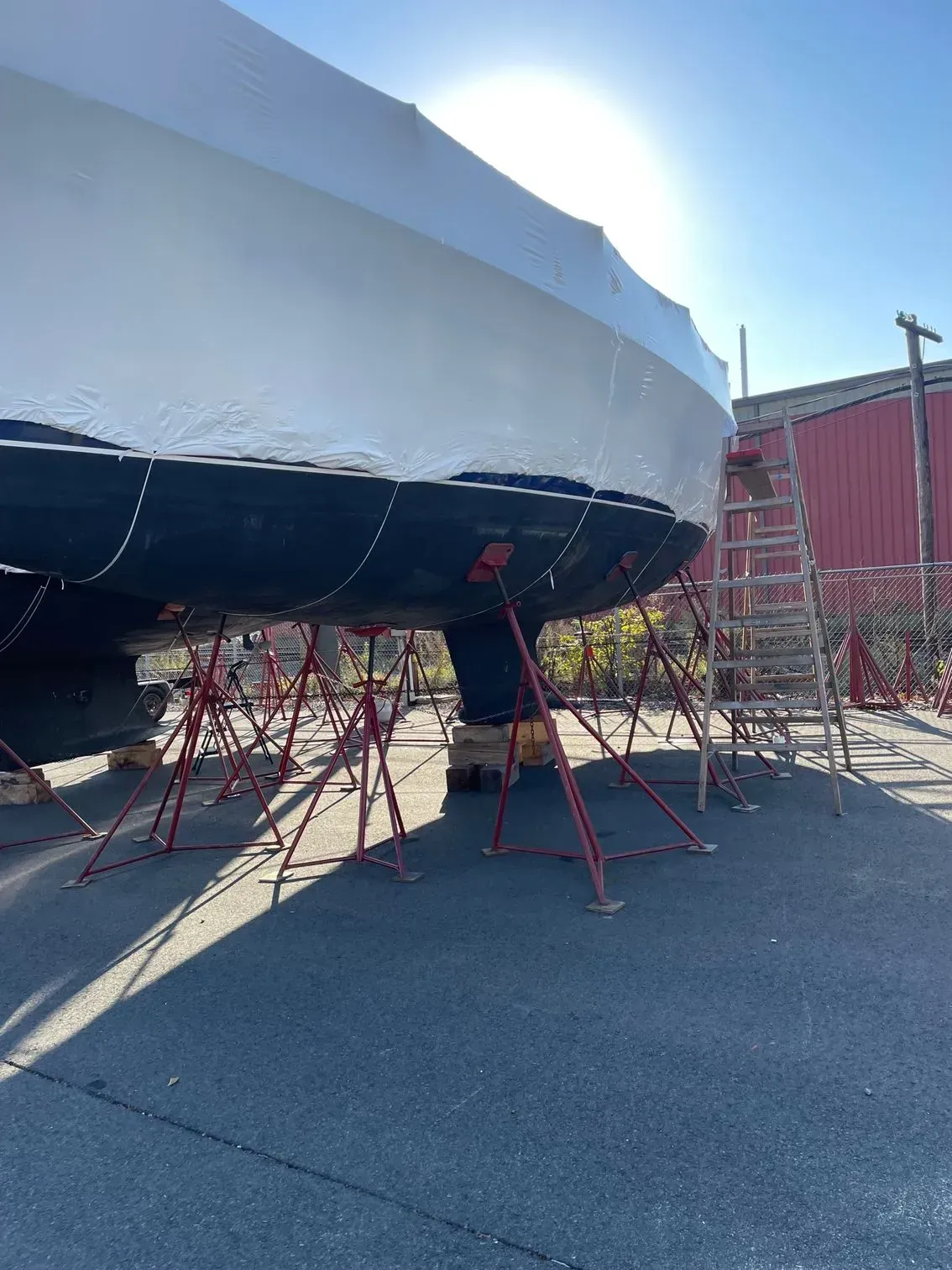 A boat hull wrapped in white plastic sitting on red metal stands in a sunny boatyard.