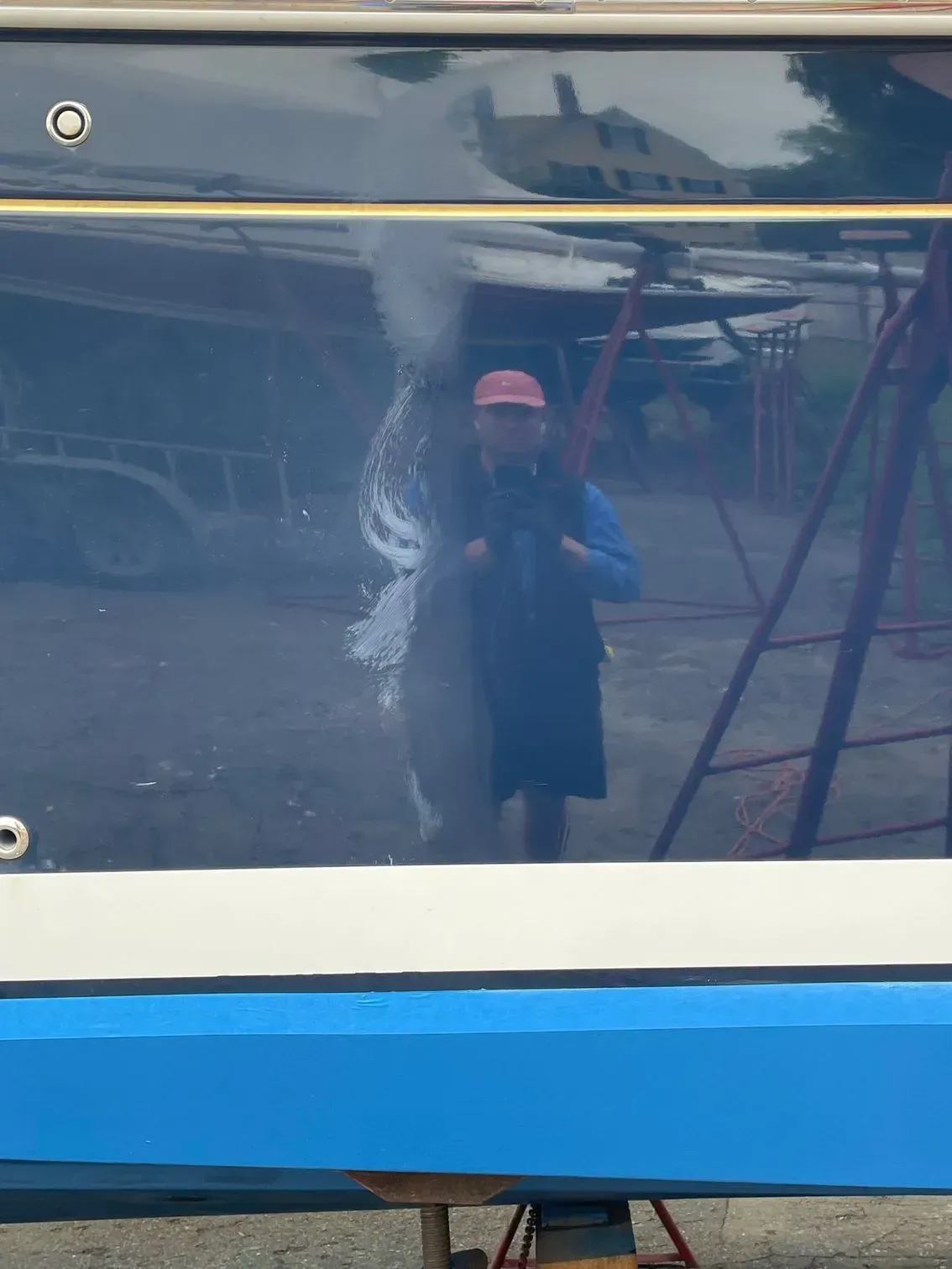A person wearing a hat and blue shirt reflected in the polished, dark hull of a boat in a yard.