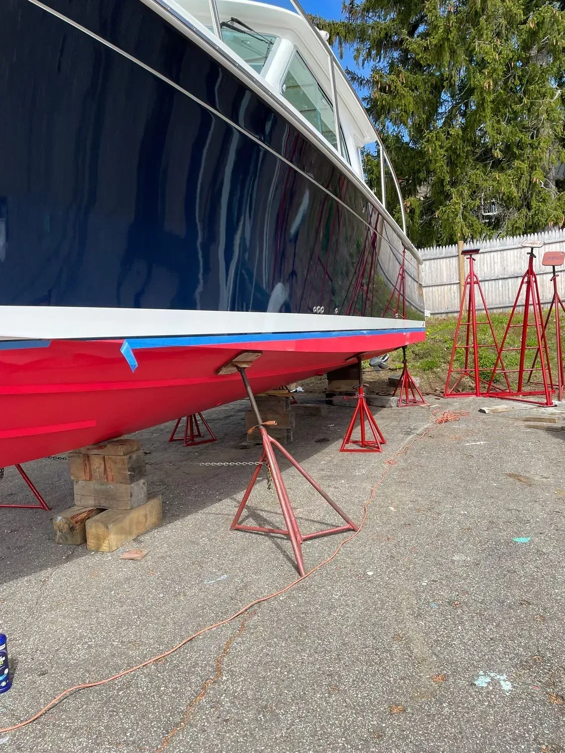 A dark blue and red boat sits on land, propped up by red metal stands in a gravel yard with a wooden fence in the back.