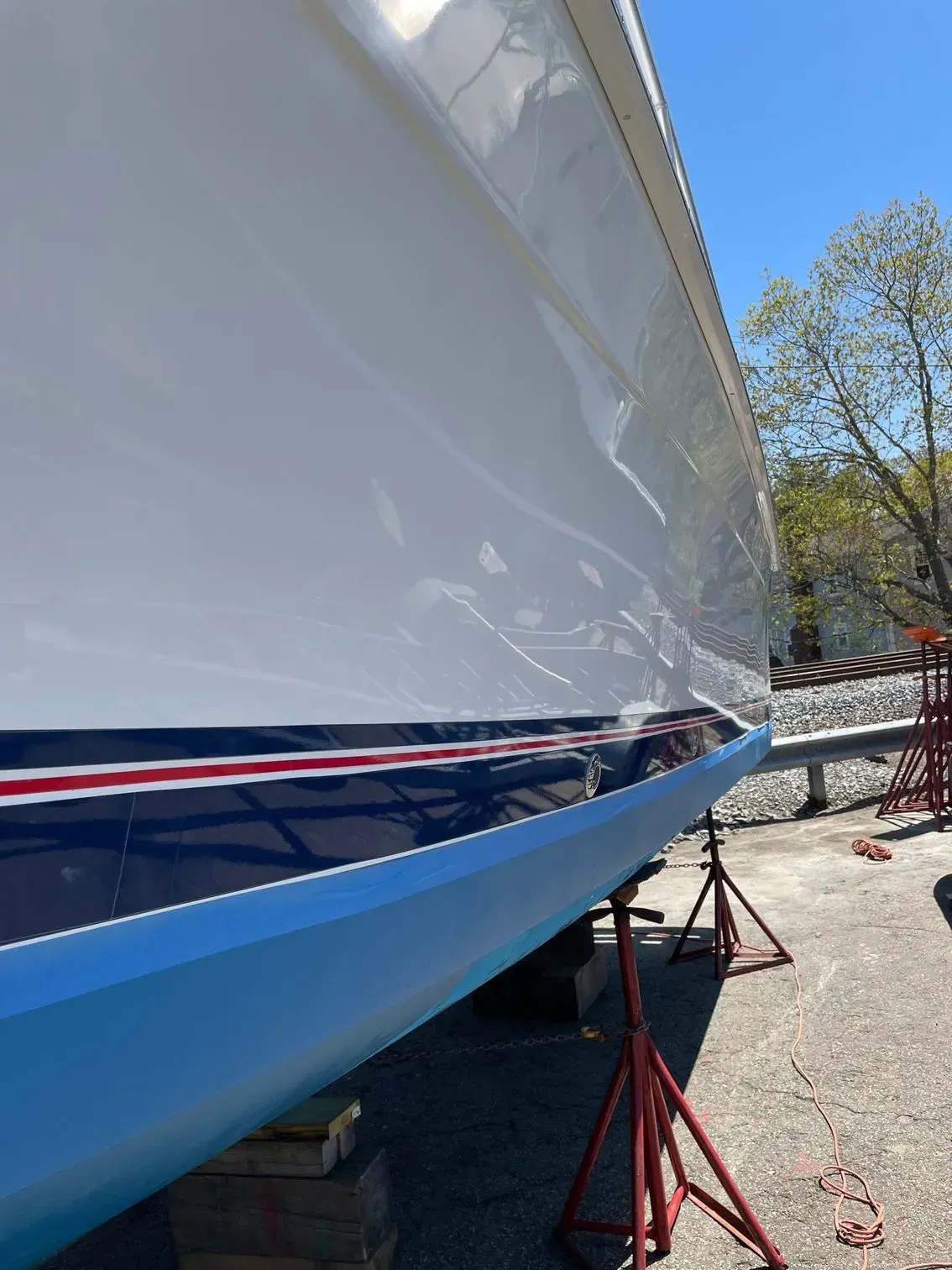 A close-up view of the white hull and blue-painted bottom of a boat on stands at a boatyard under a clear blue sky.