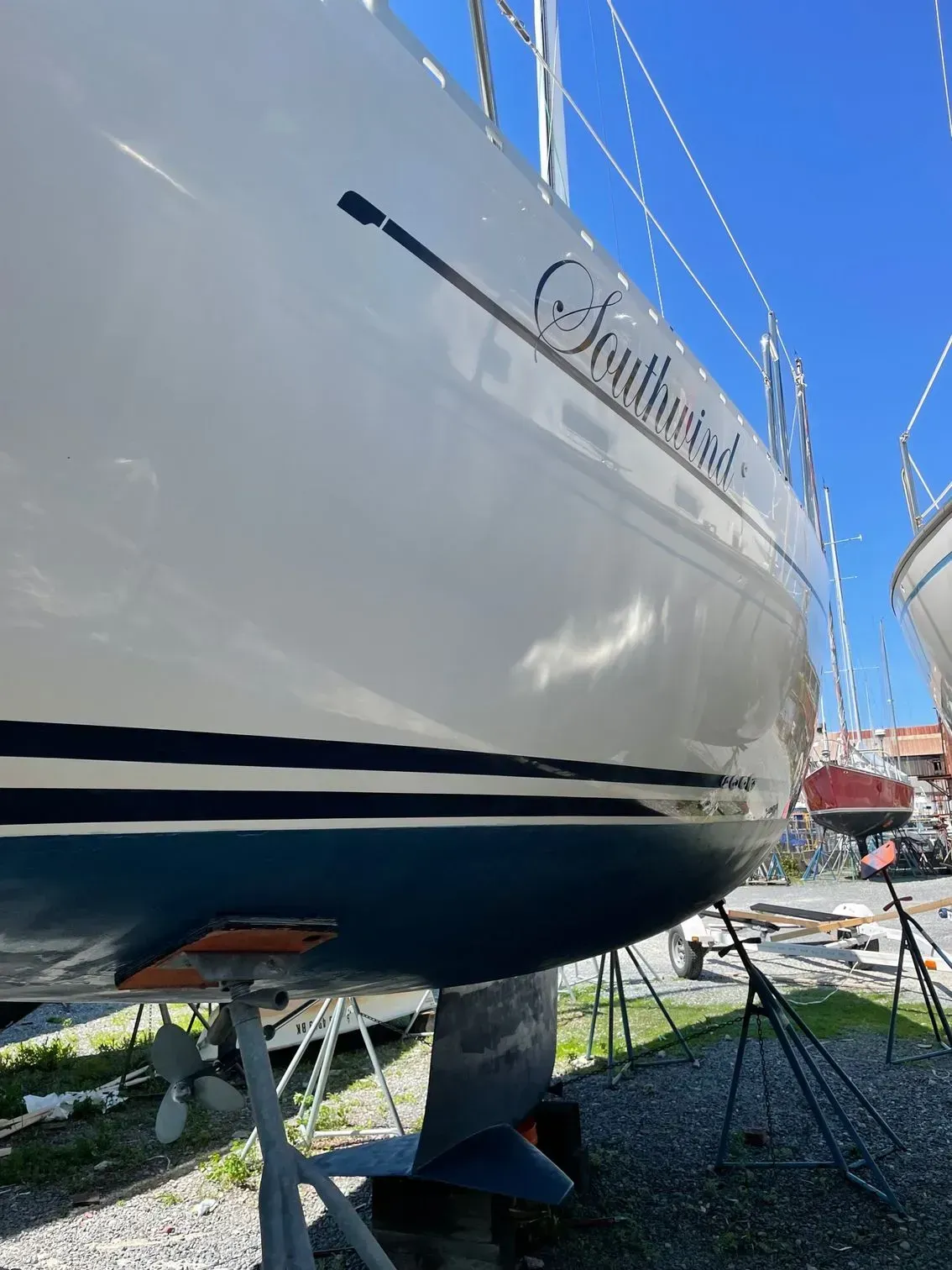 A low-angle view of a white sailboat hull with a dark blue bottom, resting on jack stands in a sunny boatyard.