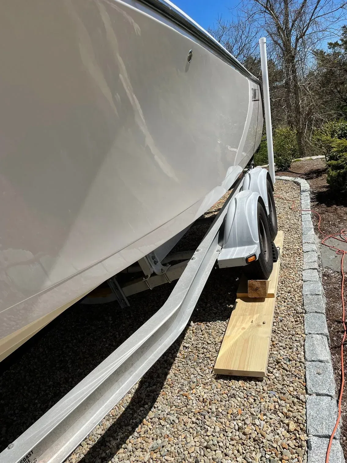 A boat trailer wheel rests on a wooden plank placed on a gravel driveway next to a stone border.