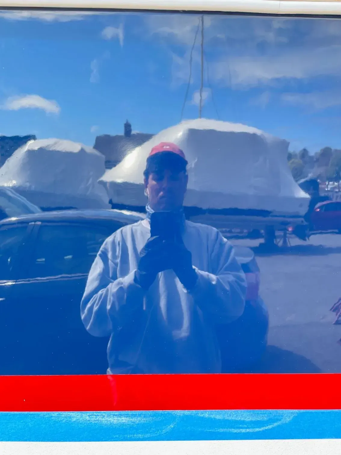 A person wearing a light-colored sweatshirt and a cap takes a selfie in a reflective blue surface at a boat yard.
