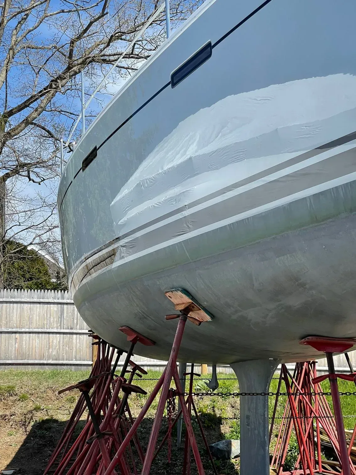 A sailboat hull sitting on red jack stands in a grassy area with a wooden fence and bare trees in the background.