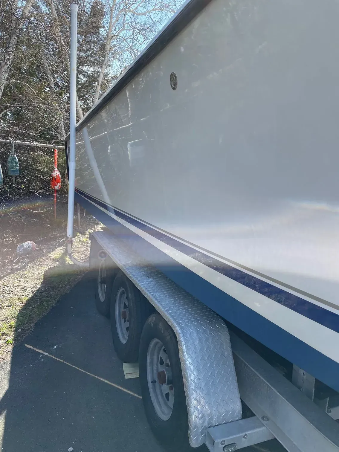 Side view of a white boat hull with a blue stripe resting on a tandem-axle trailer in a gravel lot.