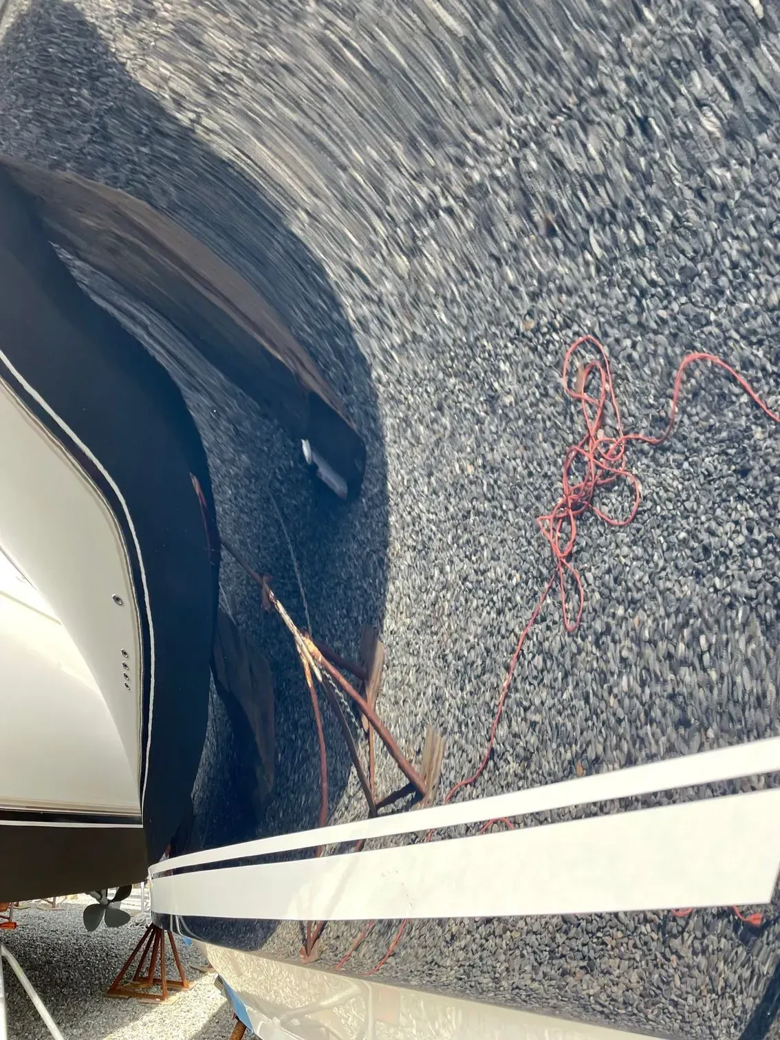 A boat on a trailer rests on a gravel surface with a red coiled rope laying on the ground nearby.