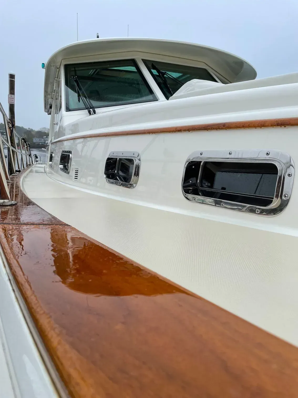 A low-angle view of a white motorboat with polished wooden trim and port windows, docked on a cloudy day.