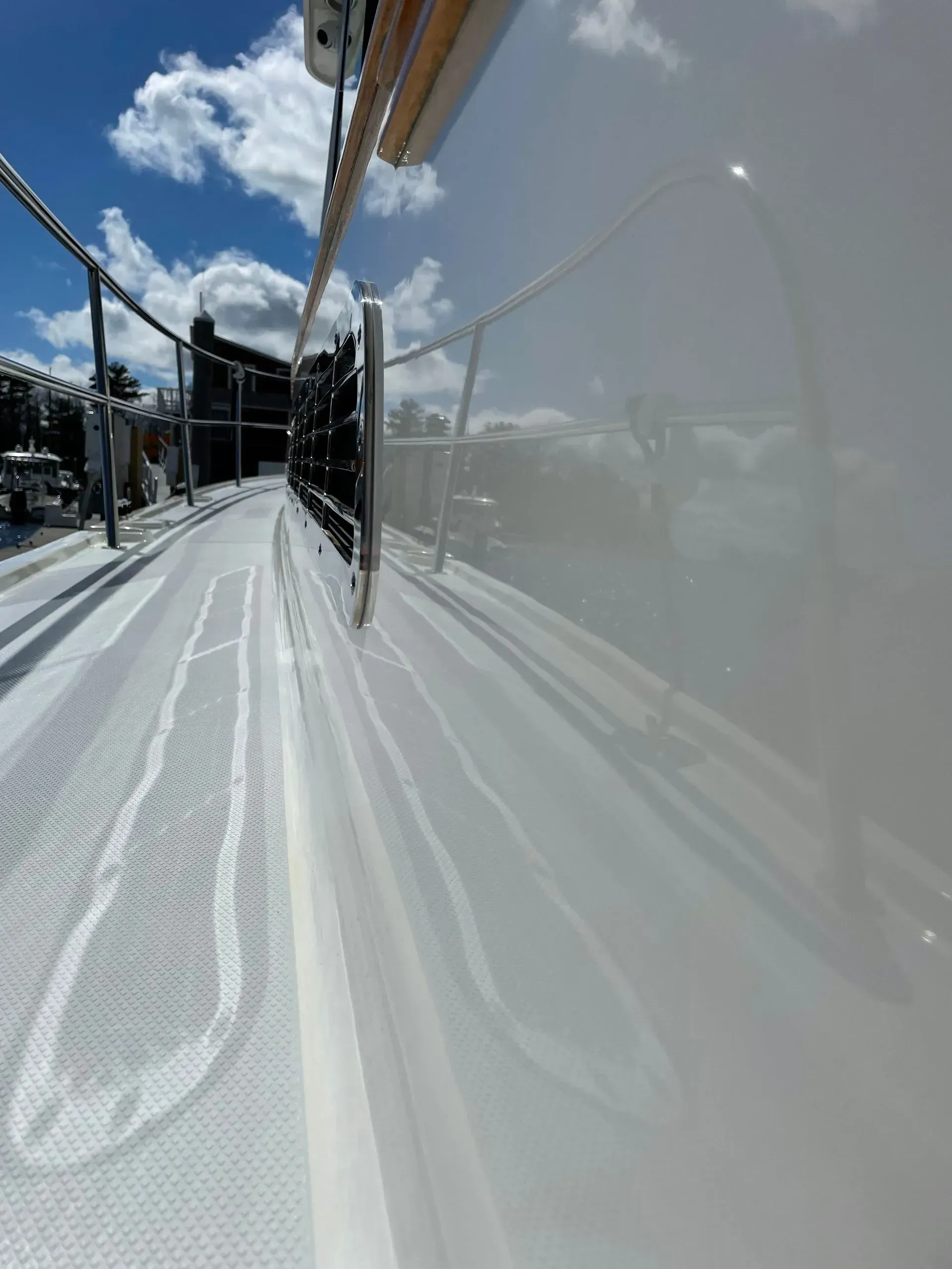 A low-angle view along the glossy white side of a boat deck, showing a metal railing, blue sky, and clouds above.