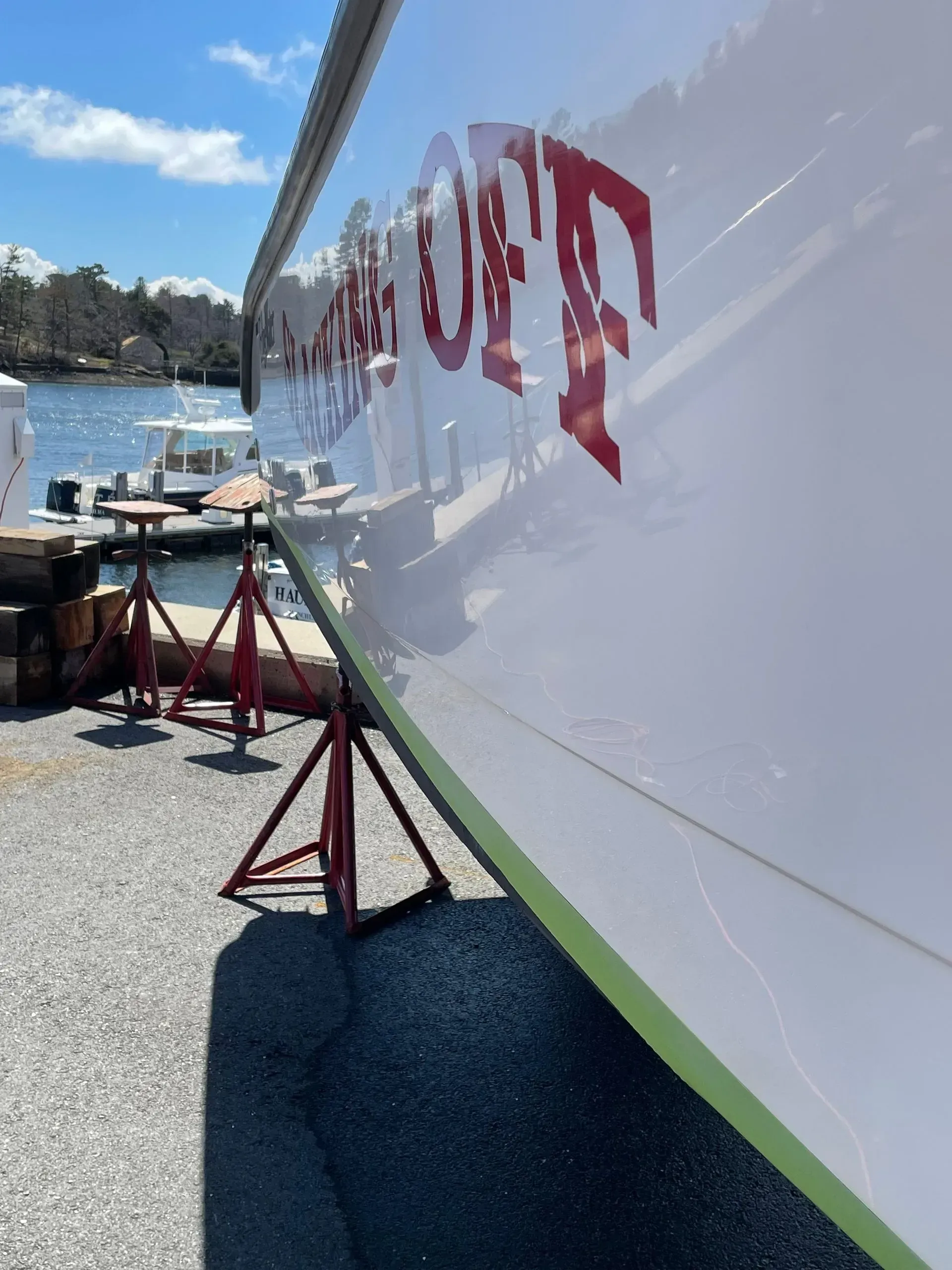 A boat hull on red metal jack stands at a marina, with the name 