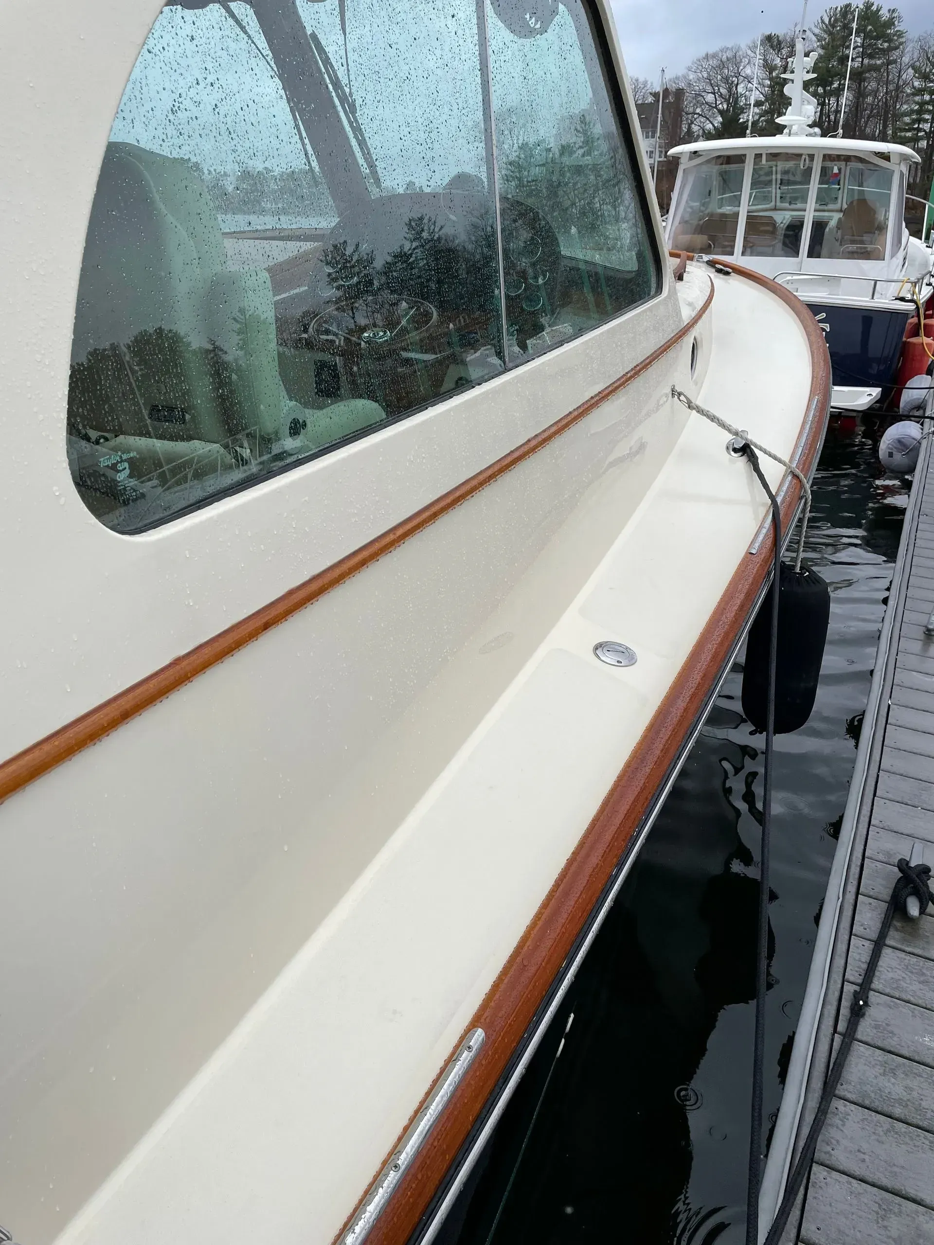 A cream-colored boat hull with wood trim docked at a pier, with another boat visible in the background.