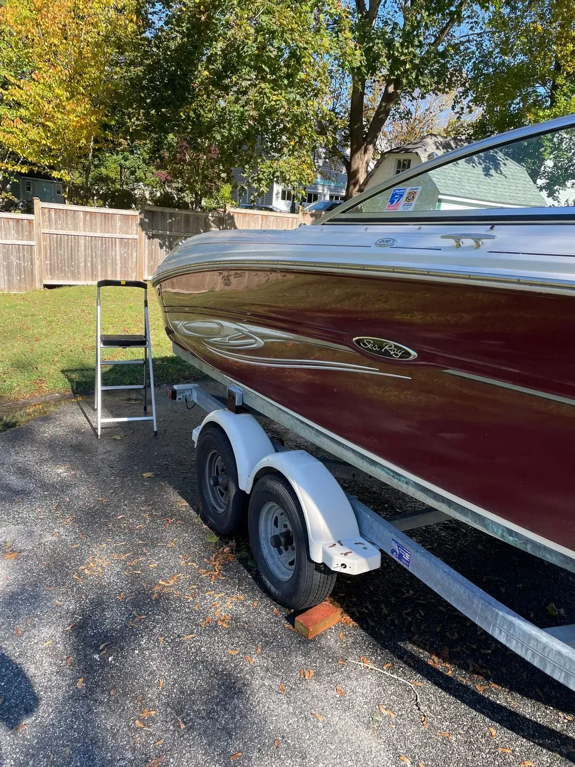 A dark red motorboat sits on a trailer in a grassy backyard, with a step ladder nearby.