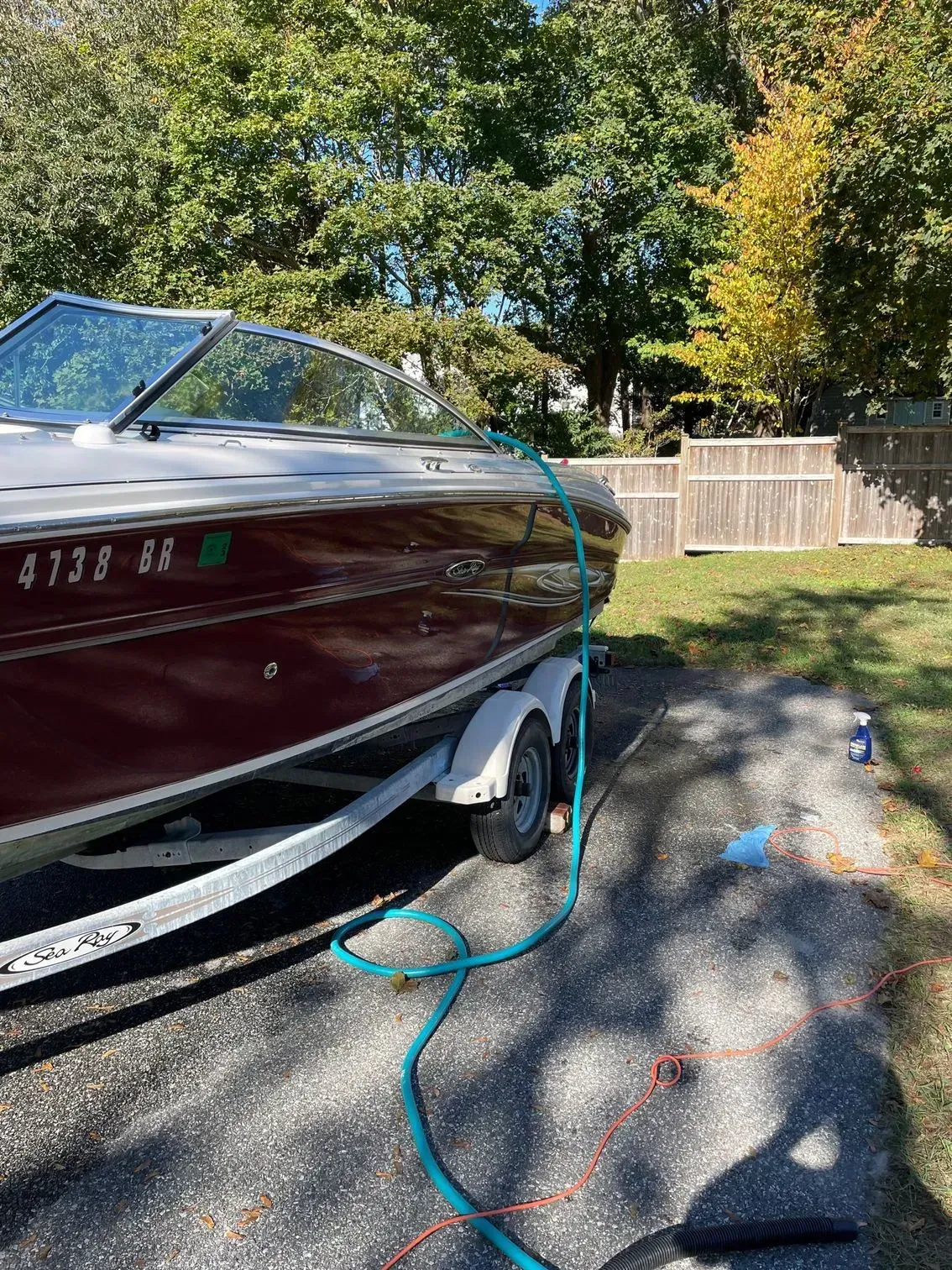A maroon motorboat on a trailer sits in a gravel driveway with a green garden hose draped over its side.